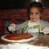 Una niña pequeña con harina en el rostro sonríe mientras esparce salsa de tomate sobre la masa de pizza
